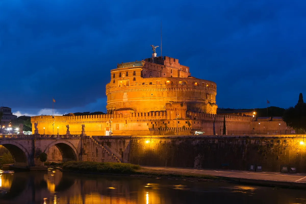 Castel Sant'Angelo lit gold at dusk beside the Tiber, a round stone fortress with a bronze angel on its summit and an arched bridge in the foreground